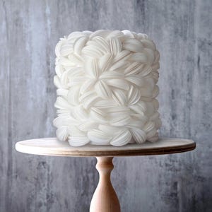 May include: A cylindrical white cake decorated with numerous overlapping petal-like layers, resting on a light-coloured wooden cake stand. The cake features a textured, layered design covering its entire exterior surface in a uniform white colour.