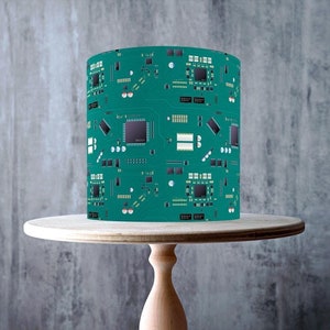 May include: A green and black circuit board cake topper. The cake topper is on a wooden cake stand. The cake topper is made of edible icing and is perfect for a tech-themed party.