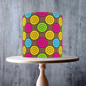 May include: A cylindrical lampshade with a brown background and a repeating pattern of colorful circles in yellow, pink, blue, and green. The lampshade sits on a light-colored wooden cake stand. The image includes a "Made in the UK" sticker and a vegan symbol.