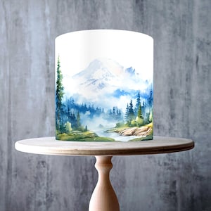 Puede incluir: Una tarta blanca con un diseño de montaña y bosque en acuarela, expuesta en un soporte para tartas de madera clara. La tarta presenta un paisaje escénico con un río, árboles y una montaña nevada. El soporte para tartas es redondo con una base de pedestal.