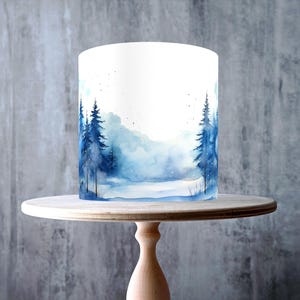 May include: A white cake with a watercolor winter forest design, displayed on a light wood cake stand. The cake features blue trees and a snowy landscape. The stand has a round top and a turned pedestal base.