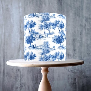 Peut inclure: Un abat-jour cylindrique avec un imprimé de scène pastorale bleu et blanc est posé sur un présentoir à gâteau en bois clair. Le présentoir a un dessus rond et un pied en forme de colonne. L'abat-jour est décoré d'arbres et de personnages.