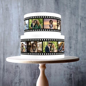 May include: A two-tiered white cake featuring a black film strip design wrapped around each tier. The film strip contains various photographic images and large numbers from two to eight. The cake is displayed on a light-colored wooden cake stand.