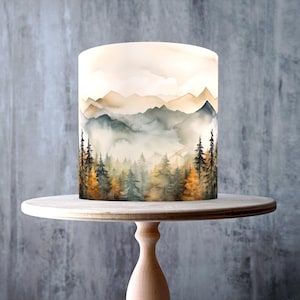 Puede incluir: Una tarta redonda con un diseño de montaña y bosque, expuesta en un soporte para tartas de madera clara. La tarta presenta un paisaje al estilo acuarela con árboles y montañas en tonos verdes, marrones y amarillos. El soporte para tartas es de madera clara.