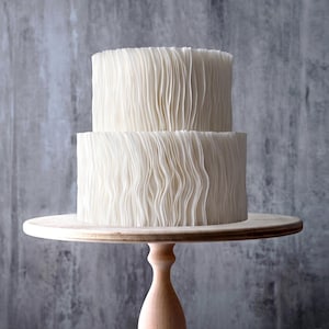 Peut inclure: Un gâteau blanc à deux étages avec une texture à volants verticaux sur un présentoir à pied en bois clair. Le gâteau présente un sommet lisse et des côtés recouverts de fines couches ondulées délicates. Deux étiquettes sont visibles.