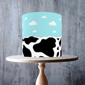 Puede incluir: Pantalla de lámpara cilíndrica con una parte superior azul cielo con nubes blancas y un patrón de piel de vaca en blanco y negro en la parte inferior. La pantalla de la lámpara se encuentra sobre un soporte para pasteles de madera clara.