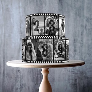 May include: A two-tiered cylindrical cake decorated with a black and white film strip design featuring photographic portraits and large numbers from 1 to 10. The cake is displayed on a light-colored wooden pedestal stand with a round base.