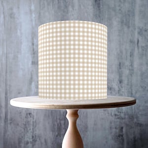May include: A light-colored wooden cake stand with a round top and a pedestal base. A cylindrical lampshade with a tan and white gingham pattern sits on top. The background is a textured gray wall.