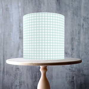 Peut inclure: Un présentoir à gâteau en bois clair avec un dessus rond et une base en piédestal. Un cylindre à motif vichy vert clair et blanc est posé dessus. Le diamètre du présentoir est d'environ 25 cm.