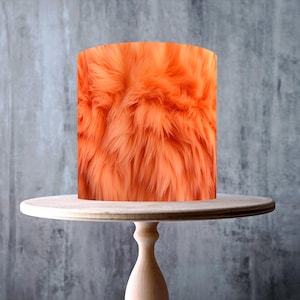 May include: An orange faux fur cake wrap sits atop a light wood cake stand. The cake wrap has a shaggy texture. The cake stand has a round top and a pedestal base. The background is a gray wall.