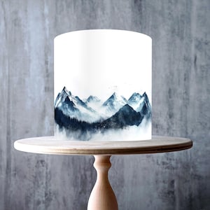 Könnte beinhalten: Ein weißer Kuchen mit einem Berglandschaftsdesign in Blau- und Grautönen, präsentiert auf einem Kuchenständer aus hellem Holz. Der Kuchenständer hat eine runde Oberseite und einen Sockel. Enthält einen "Made in the UK"-Aufkleber und einen "GMO Free, Vegan, Gluten Free, Dairy Free, Nut Free"-Aufkleber.