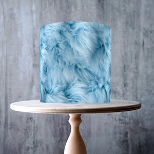 May include: A light blue cake wrap with a fluffy, fur-like texture, displayed on a light wooden cake stand. The stand has a round top and a pedestal base. The image includes a "Made in the UK" sticker and a "GMO Free, Vegan, Gluten Free, Dairy Free, Nut Free" sticker.