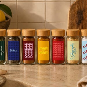 Pode incluir: Sete frascos de especiarias de vidro transparente com tampas de madeira clara, cada um com um rótulo retangular único. Os rótulos exibem os textos Origan, Poivre, Cannelle, Curry, Paprika, Gingembre e Sel em várias cores e estilos de tipografia.