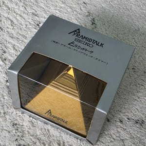 May include: A silver-colored cardboard box containing a gold-colored pyramid-shaped Seiko Pyramidtalk clock. The box features the text "PYRAMIDTALK SEIKO" and Japanese characters, with a clear plastic window displaying the gold pyramid inside.