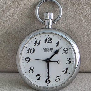 May include: A silver-tone Seiko Quartz pocket watch with a round case, a white dial, and black Arabic numerals. The watch features a top winding crown with a circular ring attachment. The dial displays the text Seiko Quartz and Japan 7550-0010 T.