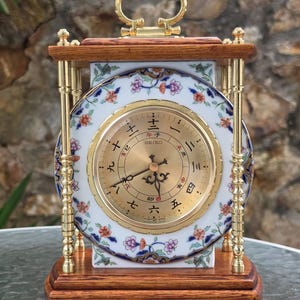 Puede incluir: Un reloj de cuarzo Seiko con esfera blanca circular decorada con motivos florales, manecillas doradas y números japoneses. El reloj está enmarcado en una estructura de madera con cuatro pilares metálicos dorados y un asa dorada superior.