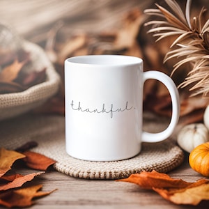 May include: White ceramic coffee mug with the word "thankful" written in black cursive script. The mug is sitting on a beige knitted coaster and surrounded by autumn leaves.
