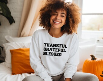Peut inclure: Un sweat-shirt blanc à manches longues avec les mots "Thankful Grateful Blessed" imprimés en noir sur le devant. Le sweat-shirt est porté par une personne aux cheveux bouclés assise sur un lit avec un oreiller blanc et orange et une citrouille.