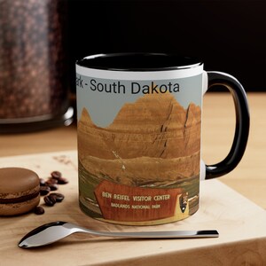 May include: White ceramic coffee mug with a black rim and handle. The mug features a scenic image of the Badlands National Park in South Dakota with the text "Ben Reifel Visitor Center, Badlands National Park" on a brown sign.