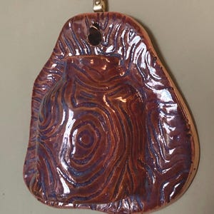 May include: A handmade ceramic wall pocket, featuring an organic shape and a textured, swirling pattern. The glaze is a mix of brown and purple hues. A small, gold-coloured metal loop is attached at the top for hanging.