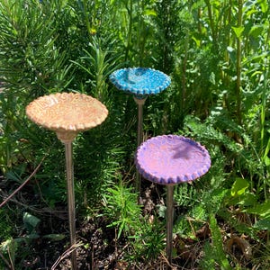 May include: Three ceramic garden stakes featuring circular, scalloped-edge tops in tan, blue, and purple. Each top displays a raised, swirling embossed pattern and is mounted on a slender, straight metal rod for insertion into the ground.