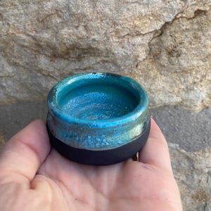 May include: A small, teal blue ceramic bowl with a black rim. The bowl is glazed and has a textured surface.