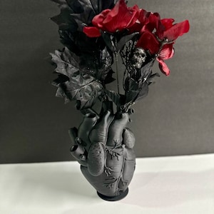 May include: A black anatomical heart-shaped vase with a bouquet of dark red flowers and black leaves. The vase has a detailed anatomical design.