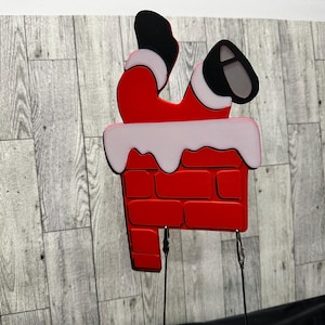 May include: A red and white Christmas decoration of Santa Claus stuck in a chimney. The chimney is made of red brick with white frosting on top. The decoration has two metal stakes for inserting into the ground.