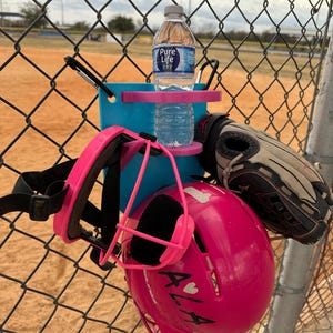 May include: A pink softball helmet, face mask, and a blue water bottle holder with a water bottle are attached to a chain-link fence. A black and tan baseball glove hangs nearby. The helmet has the letters "ALA" and a heart on it.