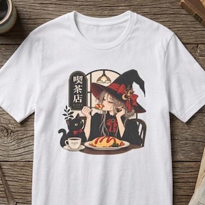 Peut inclure: Un t-shirt blanc à manches courtes avec une illustration représentant une personne portant un chapeau de sorcière, mangeant à une table avec un chat noir. Le motif inclut une tasse de café, une assiette et un panneau avec le texte japonais 喫茶店.