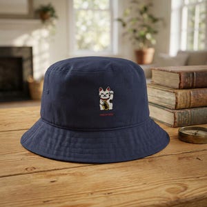 May include: A navy blue bucket hat featuring an embroidered white beckoning cat graphic on the front. The cat has a red collar and holds a gold coin. Below the cat, the text "HERE MY NECK" is embroidered in small red capital letters.