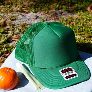 May include: A green trucker hat with a mesh back and a white label that says "OTTO WORLD FAMOUS FOAM FRONT TRUCKER". The hat is sitting on a light blue towel with a tube of sunscreen and an orange.