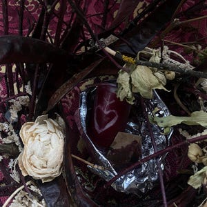May include: A still life arrangement featuring a deep red candle with a heart design, surrounded by dried flowers, dark branches, and a patterned fabric background. The candle is partially wrapped in foil.