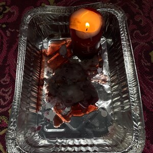 May include: A burning red candle in a silver foil tray. Melted wax and a dark, reddish residue surround the candle. The tray rests on a patterned, maroon surface.