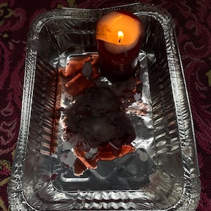 May include: A lit red candle sits in a silver foil tray, with melted wax and residue. The candle flame glows brightly, casting a warm light. The tray is on a patterned surface.