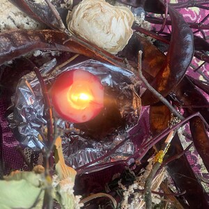 May include: A lit red candle with a bright yellow flame, surrounded by dried seed pods, twigs, and a cream-colored flower. The candle sits in a foil wrapper, with a pink and purple patterned background.