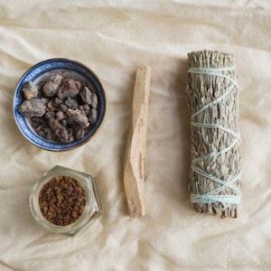 May include: A white bowl with blue trim filled with dark brown and red resin pieces, a small glass jar with brown crystals, a light brown stick, and a bundle of dried sage tied with white string.
