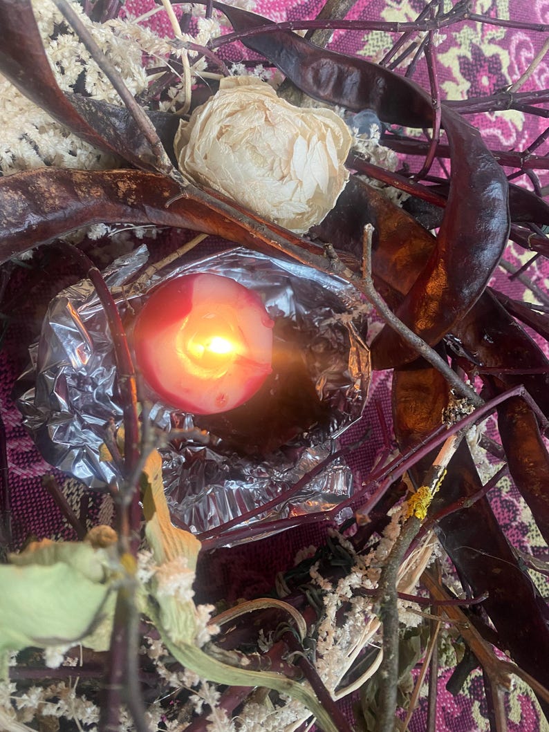 May include: A lit red candle with a bright yellow flame, surrounded by dried seed pods, twigs, and a cream-colored flower. The candle sits in a foil wrapper, with a pink and purple patterned background.