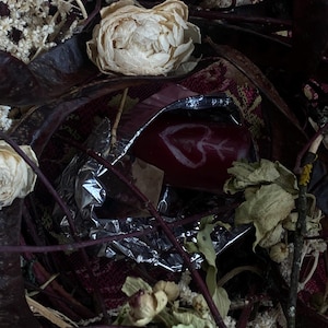 May include: A close-up of a red object with a white heart and arrow design, partially wrapped in silver foil. The image is surrounded by dried flowers, leaves, and twigs, with a patterned red and burgundy fabric in the background. The overall aesthetic is rustic and natural.