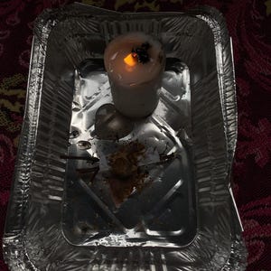 May include: A lit white candle with a dark top sits in a small, disposable aluminium foil tray. The candle's flame glows brightly, casting a warm light. The tray contains remnants of the candle and other debris.