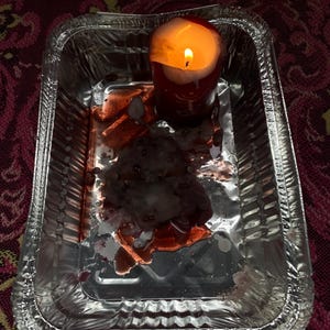 May include: A lit red candle in a silver foil tray. The candle has melted wax and is surrounded by a dark, reddish substance. The tray sits on a patterned, maroon surface.