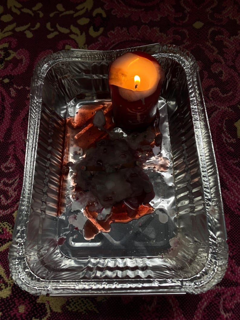 May include: A lit red candle in a silver foil tray. The candle has melted wax and is surrounded by a dark, reddish substance. The tray sits on a patterned, maroon surface.