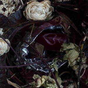 May include: A close-up shot of a red object with a white heart and arrow design, partially wrapped in silver foil. The image is surrounded by dried flowers, leaves, and twigs, with a patterned red and burgundy fabric in the background. The overall aesthetic is rustic and natural.