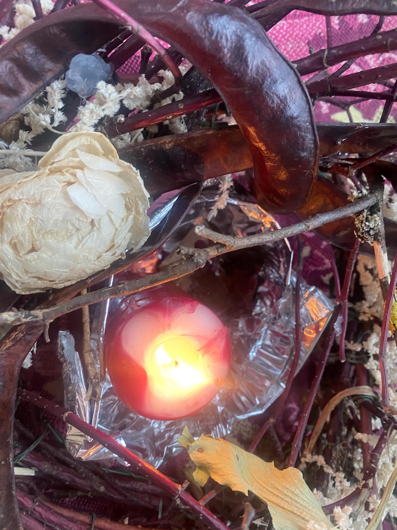 May include: A lit red candle with a yellow flame, surrounded by dried branches, seed pods, and a cream-colored flower. The background features a pink floral pattern and silver foil.
