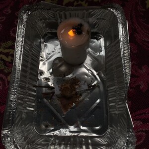 May include: A lit white candle with a dark top sits in a small, disposable aluminum foil tray. The candle's flame glows brightly, casting a warm light. The tray contains some debris from the candle.