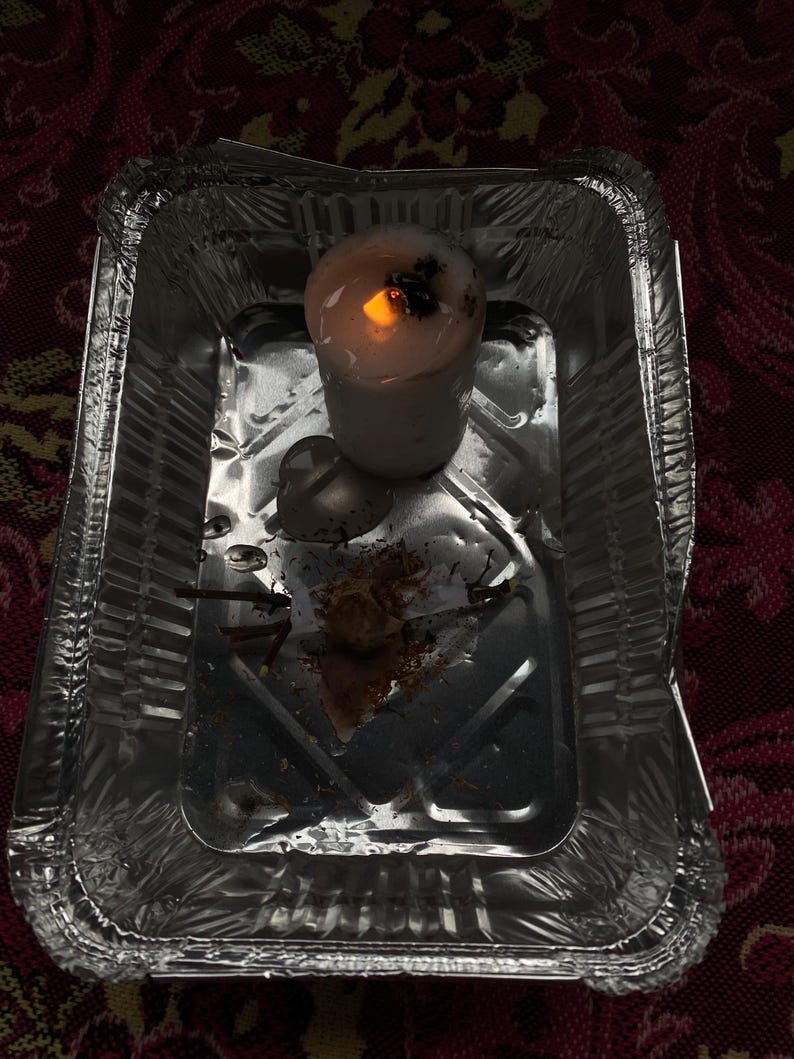 May include: A lit white candle with a dark top sits in a small, disposable aluminum foil tray. The candle's flame glows brightly, casting a warm light. The tray contains some debris from the candle.
