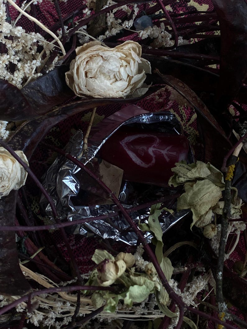 May include: A close-up shot of a red object with a white heart and arrow design, partially wrapped in silver foil. The image is surrounded by dried flowers, leaves, and twigs, with a patterned red and burgundy fabric in the background. The overall aesthetic is rustic and natural.