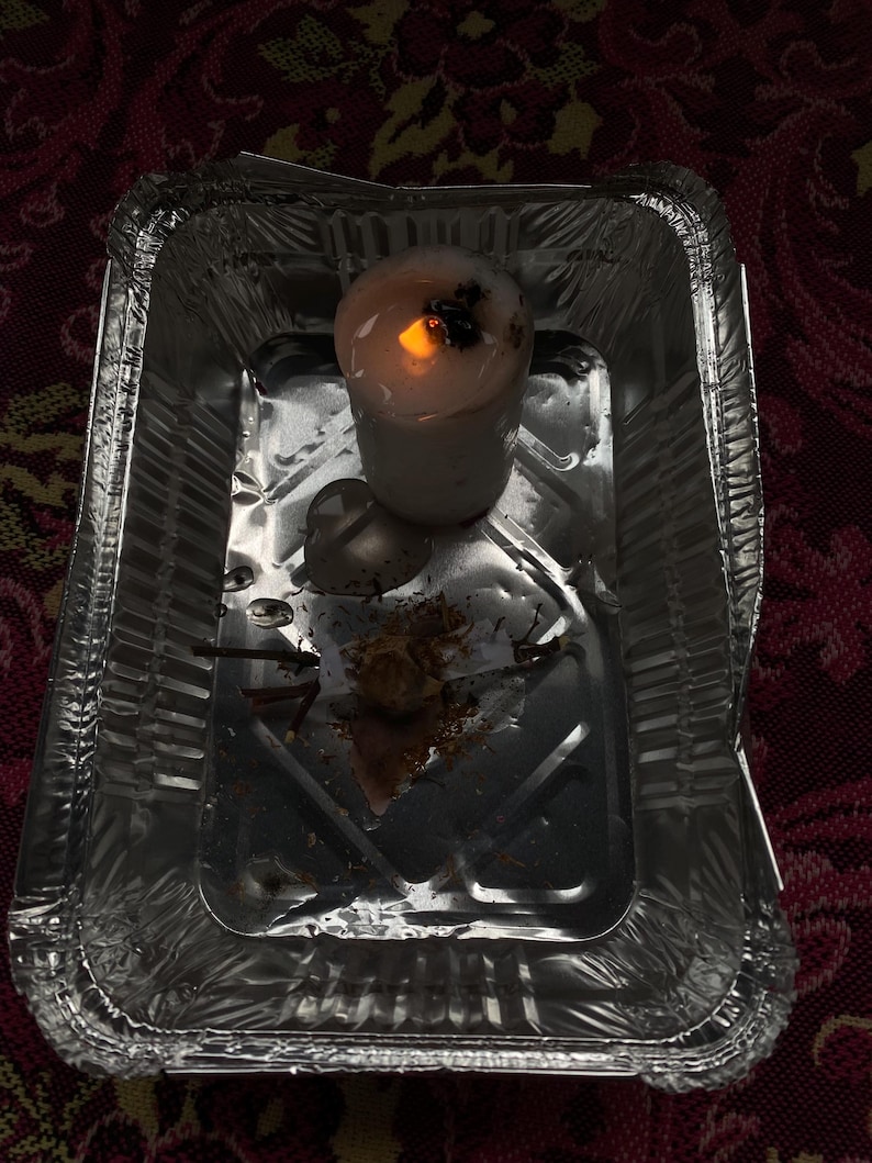 May include: A lit white candle with a dark top sits in a small, disposable aluminum foil tray. The candle's flame glows brightly, casting a warm light. The tray contains remnants of the candle and other debris.