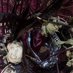 May include: A still life arrangement featuring a deep red candle with a heart design, surrounded by dried flowers, dark branches, and a patterned fabric background. The candle is partially wrapped in foil.