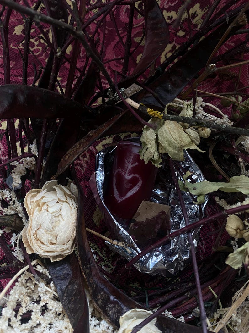 May include: A still life arrangement featuring a deep red candle with a heart design, surrounded by dried flowers, dark branches, and a patterned fabric background. The candle is partially wrapped in foil.